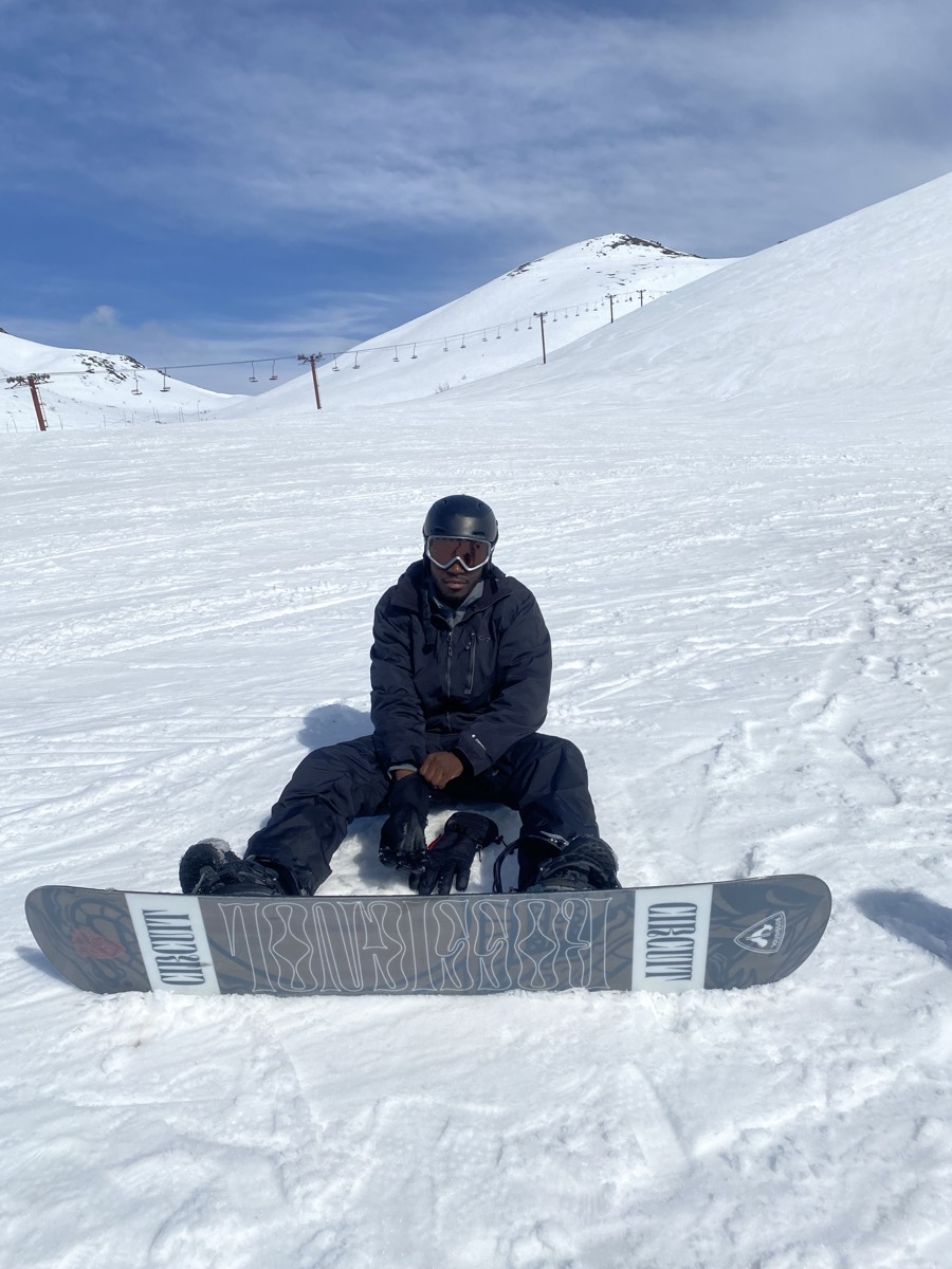 Amaya's husband seated on the snow with a snowboard on an Alaskan slope under a bright blue sky.